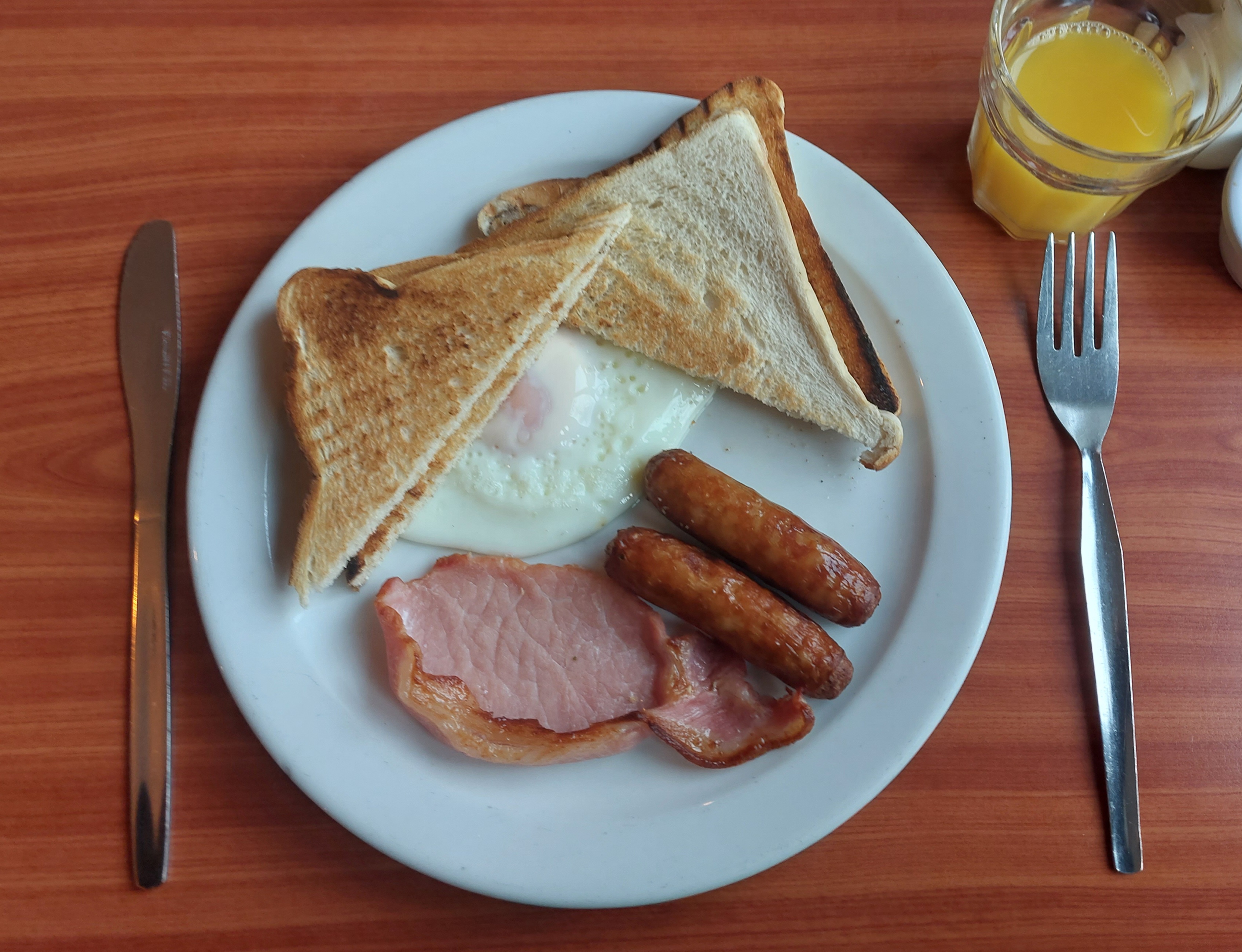 A plate full of food, including toast, eggs, sausages, and rashers with a glass of fresh orange juice to the right.